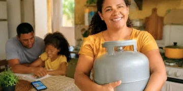 Mulher sorrindo segura botijão de gás em cozinha simples, com família ao fundo e celular sobre a mesa.